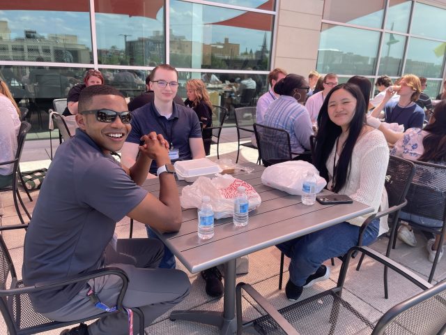 Three students sit around a table outside with to-go boxes and water in front of them. They are smiling at the camera.